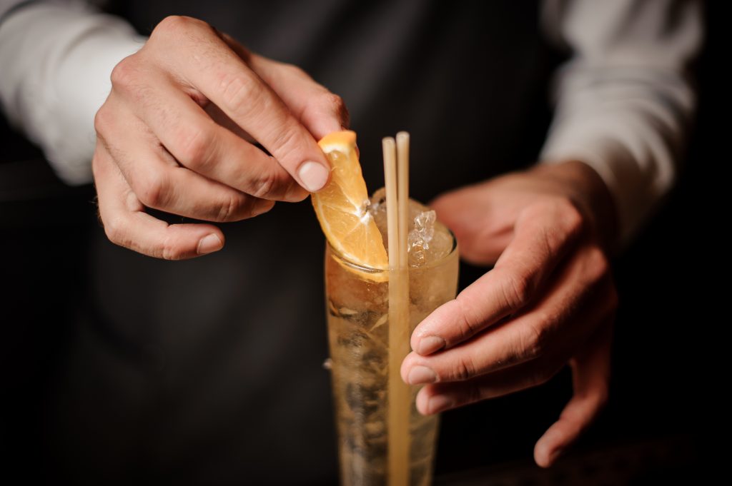 Barman decorating sweet and fresh summer cocktail with a slice of fresh orange and wooden straw